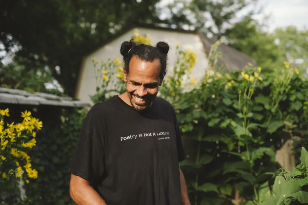 Photo of Ross Gay, poet & author, standing in front of a line of sunflowers