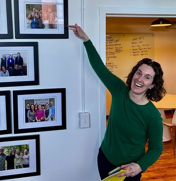 Photo of Gretchen pointing to wall of framed photos of Rowland Fellow cohorts